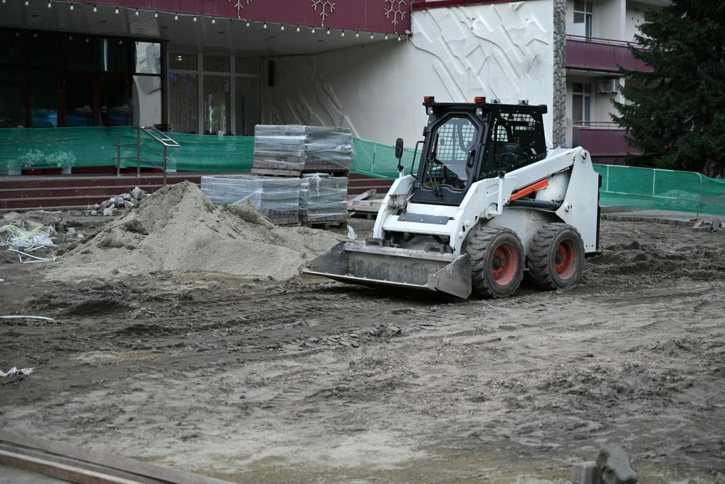 A small tractor works in the sand, filling up a hole. Shovels are on the ground. View from above.