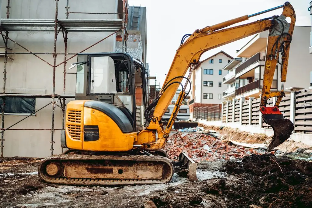 Yellow mini excavator digging fence foundation between construction site buildings